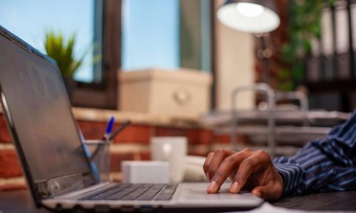 Selective focus of african american person scrolling on touchpad in brick wall workspace. Closeup of laptop at office desk being used by black male individual, updating business project presentation.